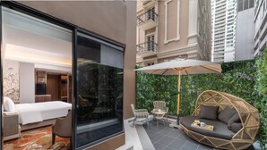 Una camera d'albergo con un letto, una parete divisoria in vetro e vista su un balcone con arredi da esterno e piante.