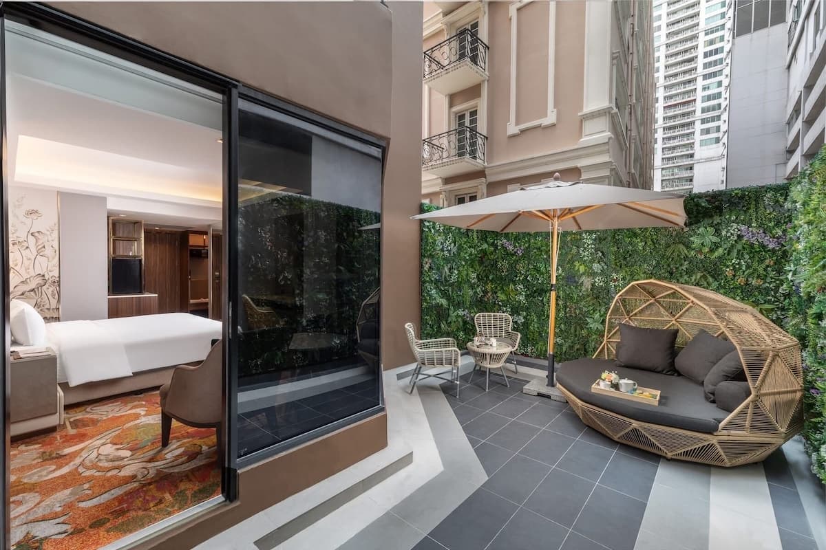 A hotel room with a bed, a glass partition, and a view of a balcony with outdoor seating and greenery.