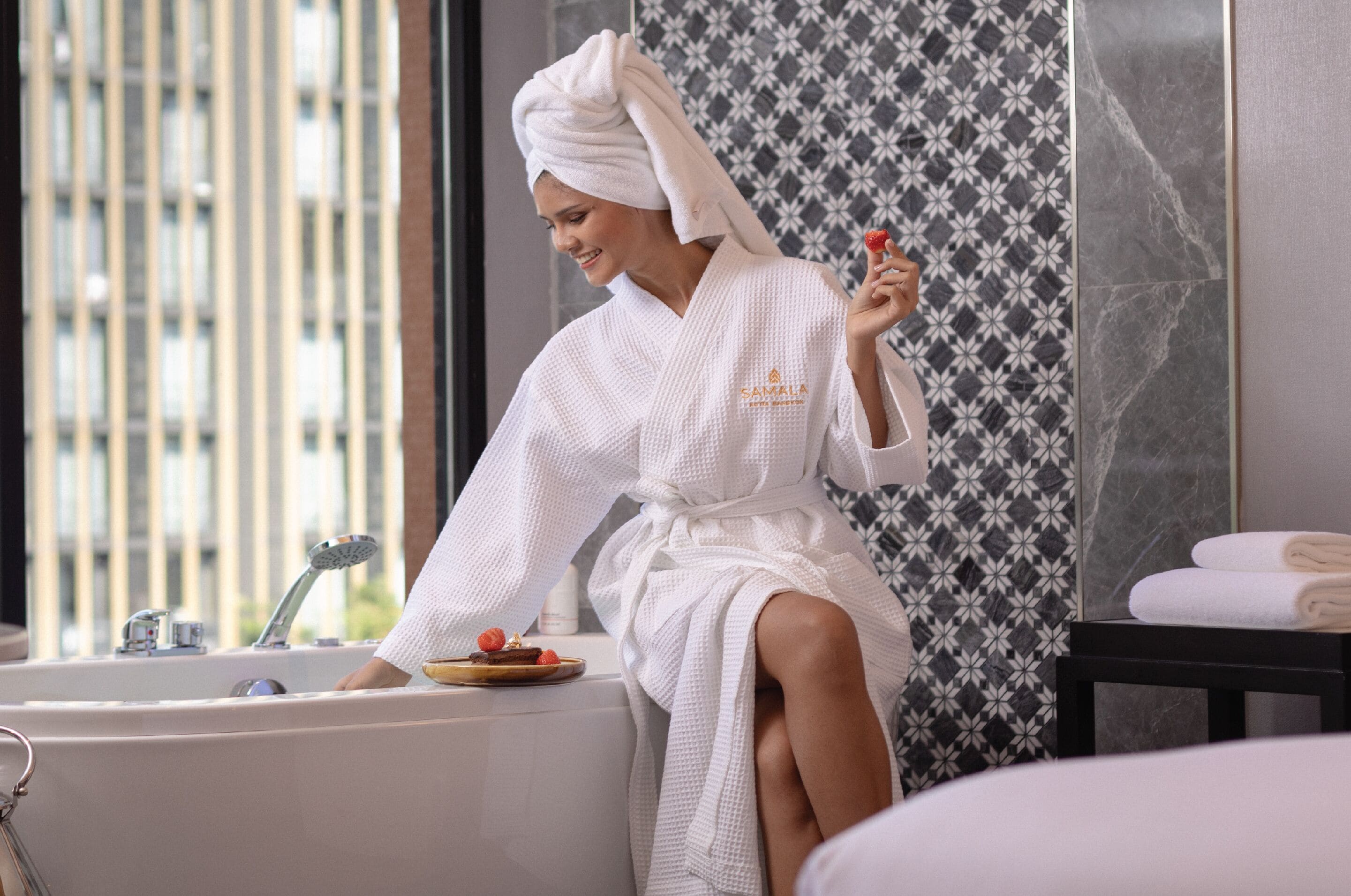 A person in a white bathrobe sitting in a bathtub with a towel and a fruit.
