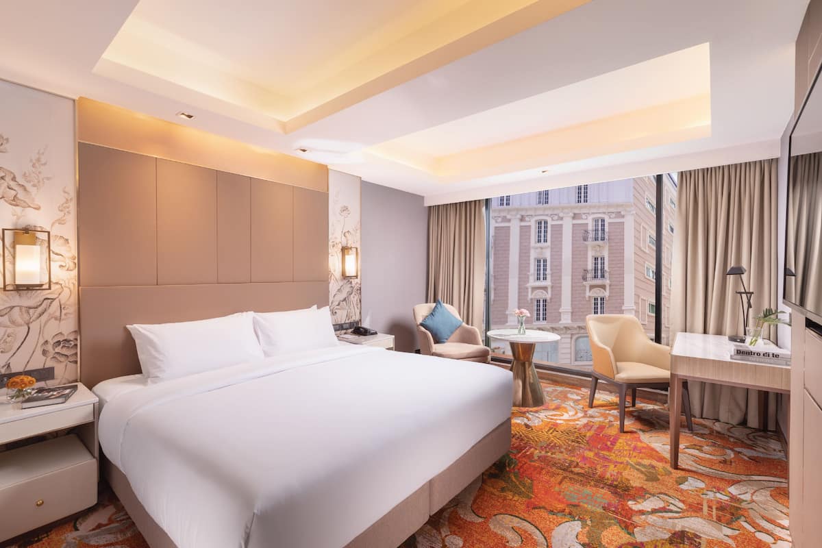 A hotel room with a large bed, a desk, a chair, and a view of a cityscape through the window.