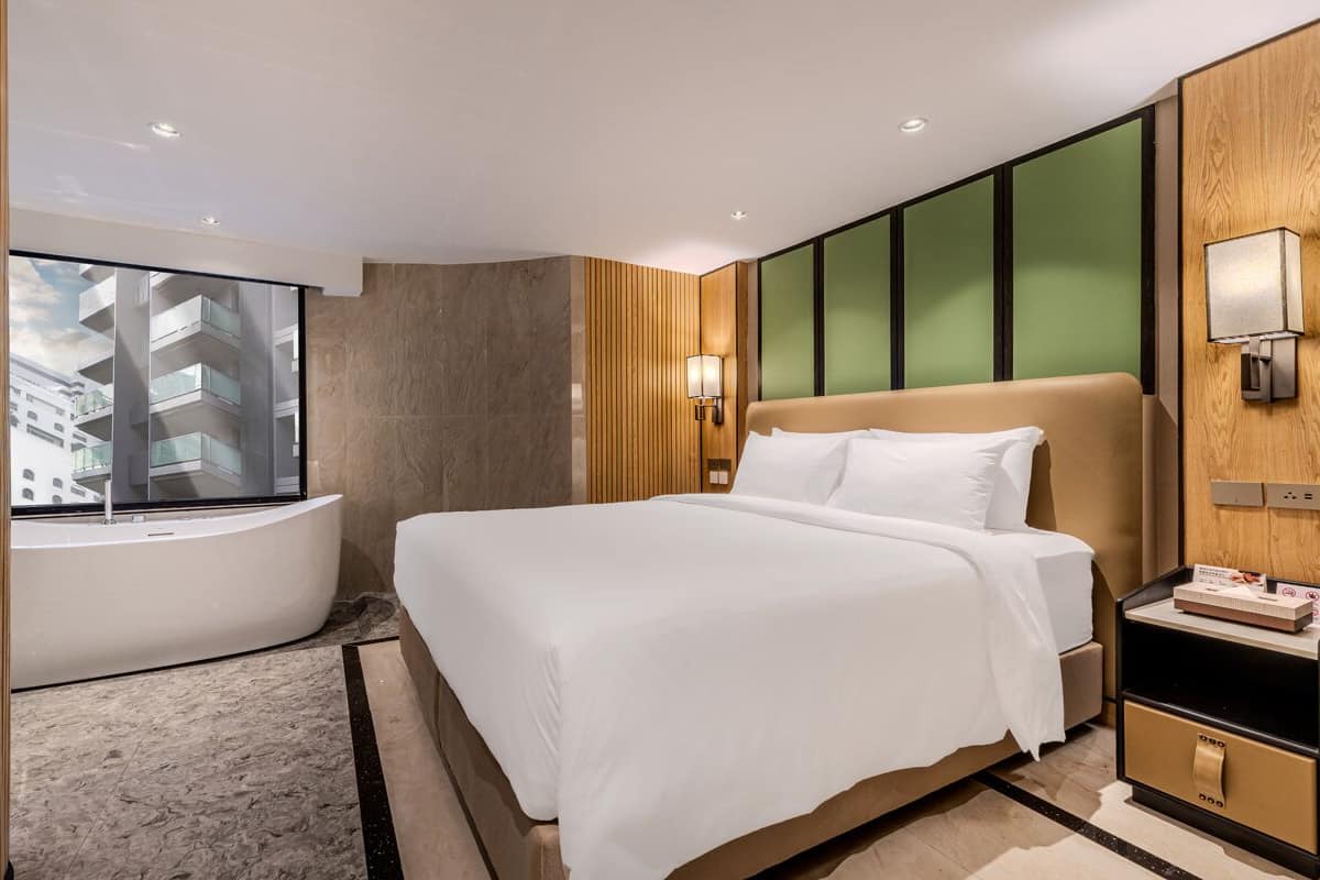 A modern hotel room with a large bed, a bedside table, a wall-mounted lamp, and a view of a building through a large window.