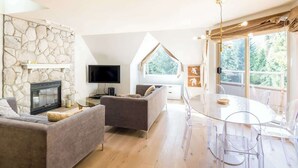 A modern living room with a stone wall, fireplace, flat-screen TV, grey sofa, and a dining area with a round table and chairs. - Ski in, Free Shuttle, Walk to Village (Whistler)