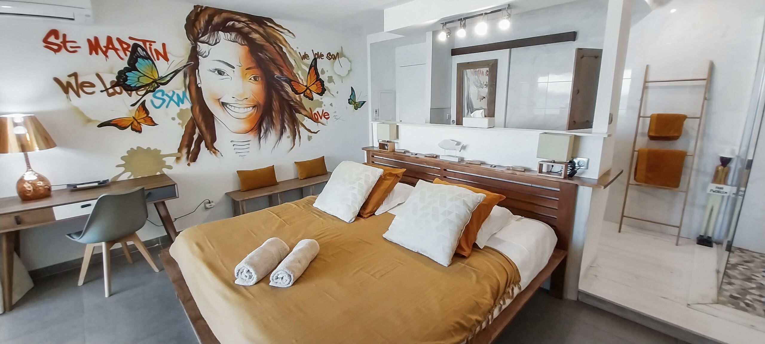 A modern hotel room with a large bed, a desk, and a chair. There is a wall mural with text and butterflies.