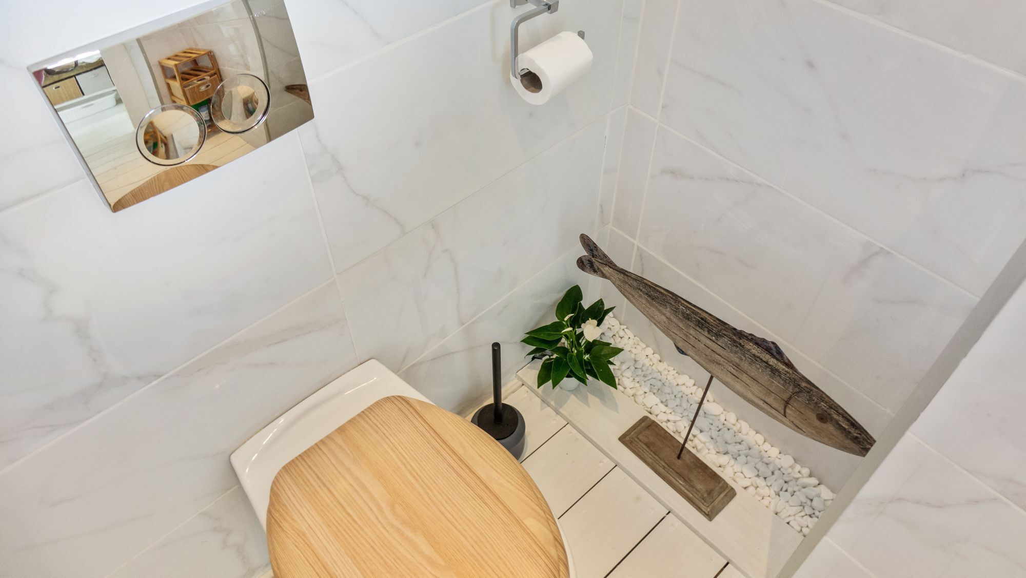 Een badkamer met een houten toiletbril, een toiletrolhouder, een toiletborstel en een plant met witte bloemen.