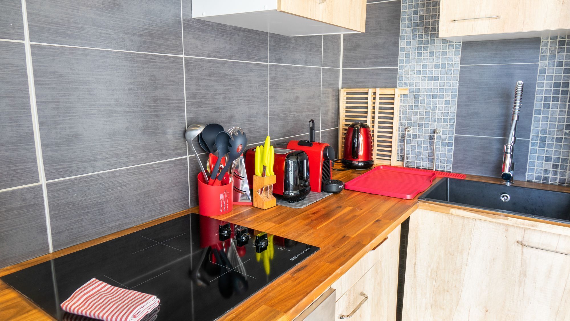 Uma cozinha moderna com azulejos cinza, um fogão por indução preto, uma torradeira vermelha, uma chaleira e uma pia.
