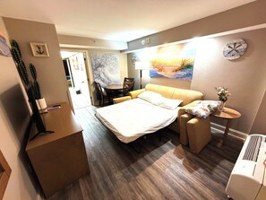 Une chambre d’hôtel avec un lit, une petite table, une chaise, un bureau, une plante en pot et une horloge murale.