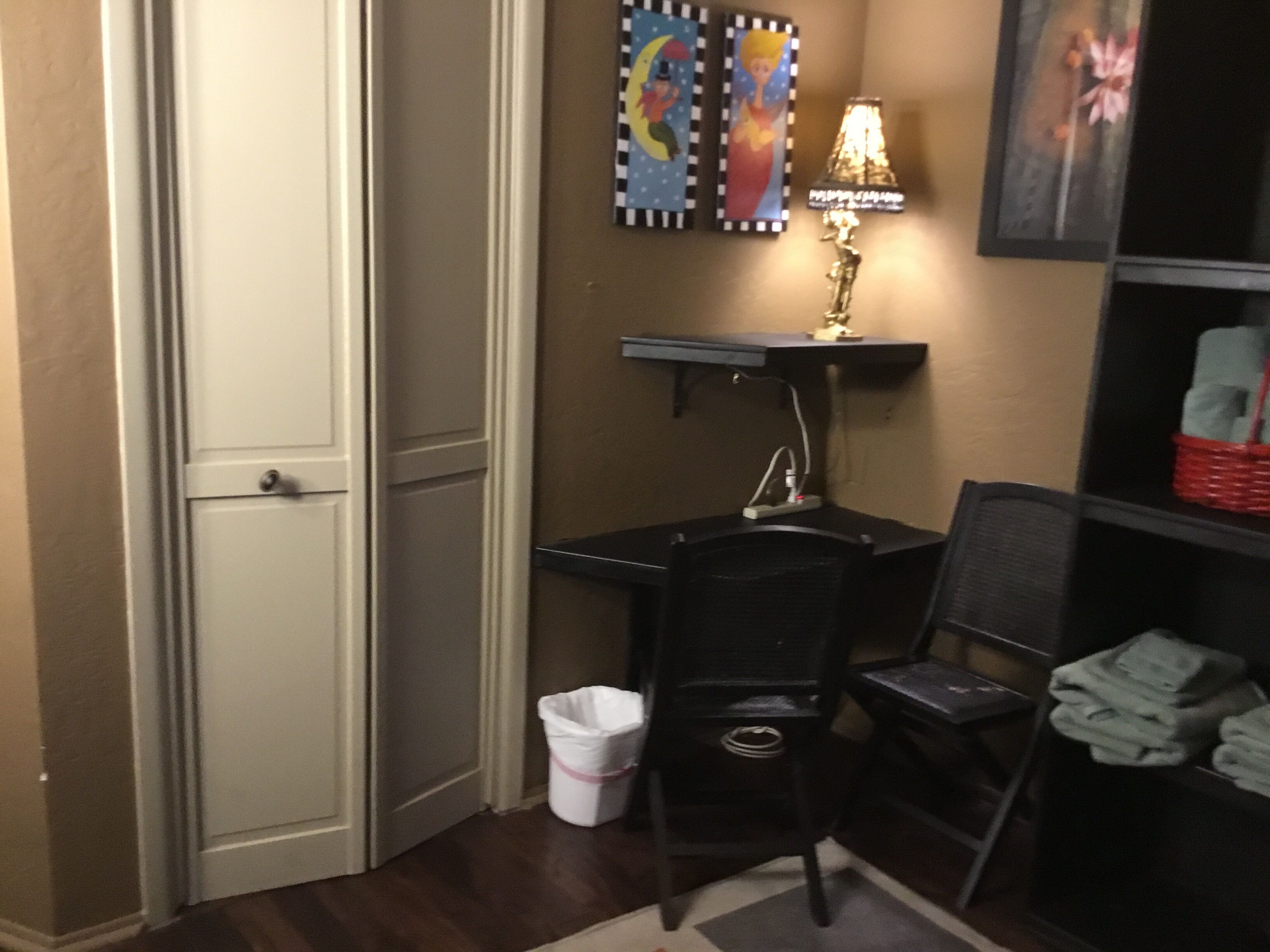 A small office area with a desk, chair, and a trash bin. There are wall decorations and a lamp on a shelf.