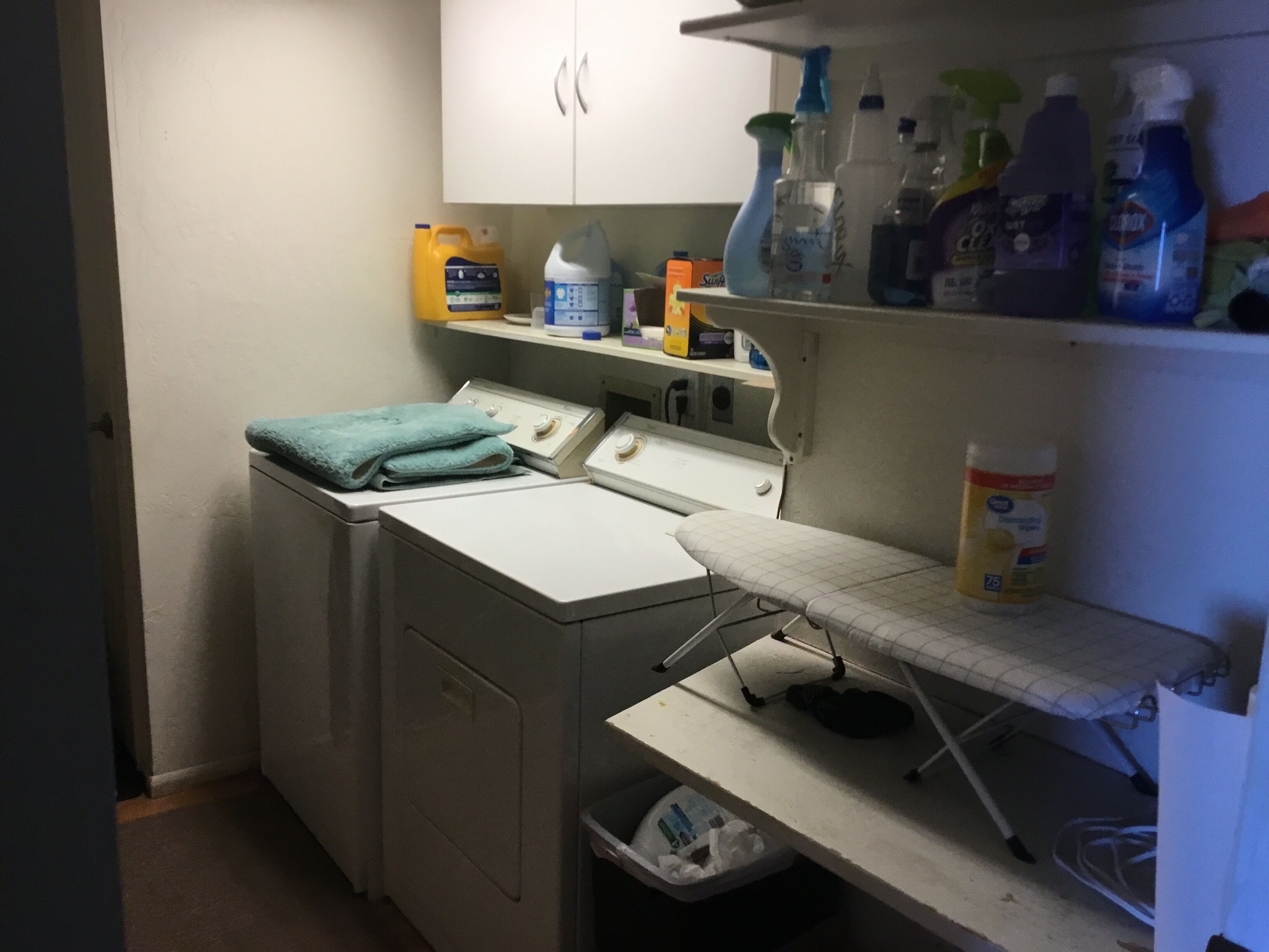Ett tvättrum med tvättmaskin, torktumlare, strykbräda och hyllor med rengöringsmedel.