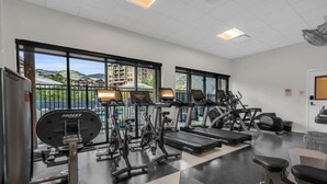 Una palestra con diverse attrezzature per esercizi, ampie finestre e vista su edifici e colline.