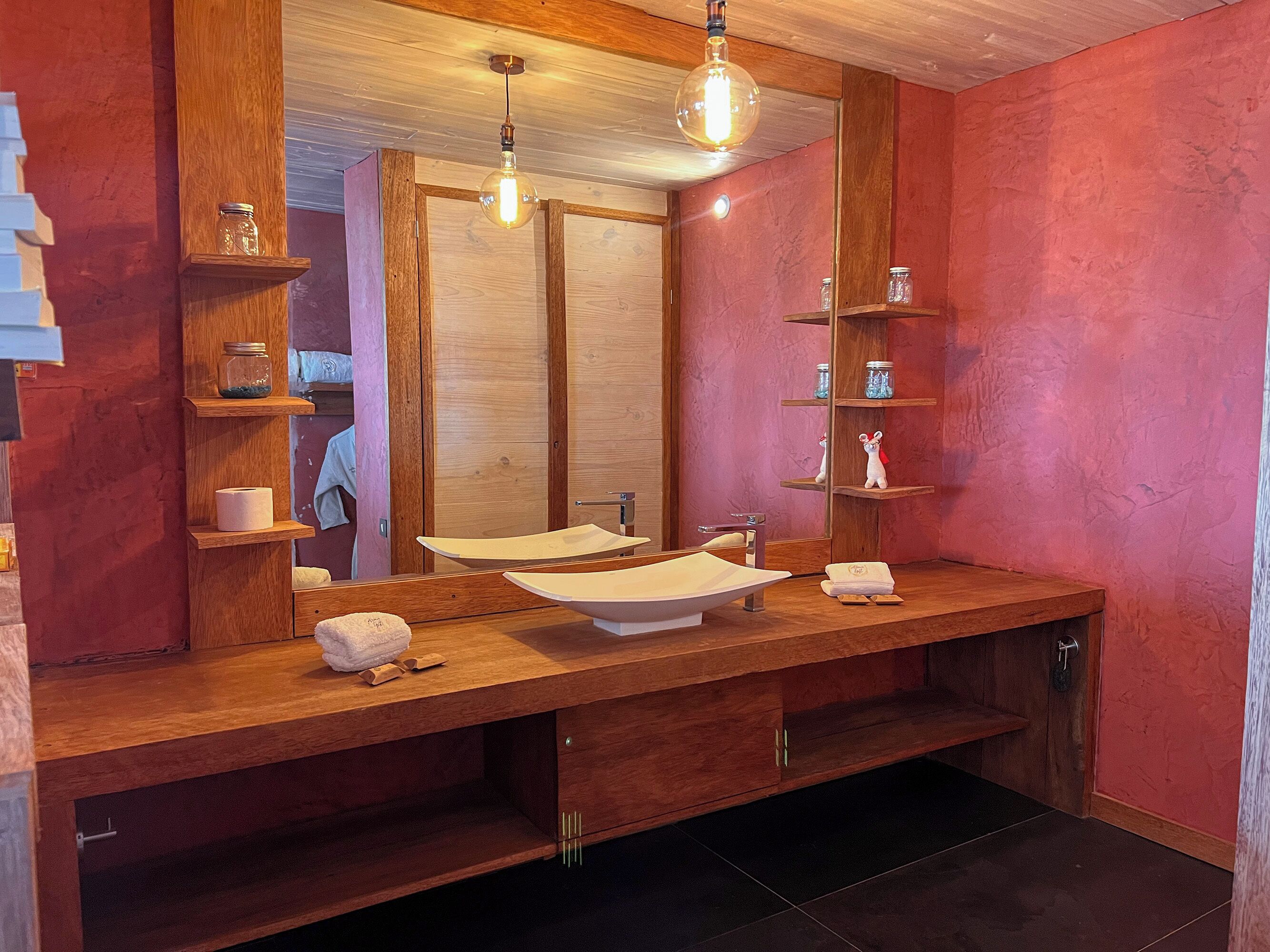 Un baño moderno con un tocador de madera, un espejo grande y un lavamanos. Hay estantes con botes y artículos de tocador.