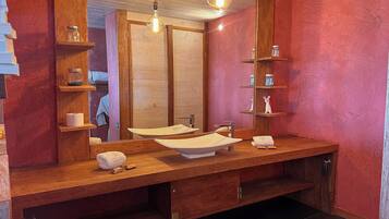 Un baño moderno con un tocador de madera, un espejo grande y un lavamanos. Hay estantes con botes y artículos de tocador.