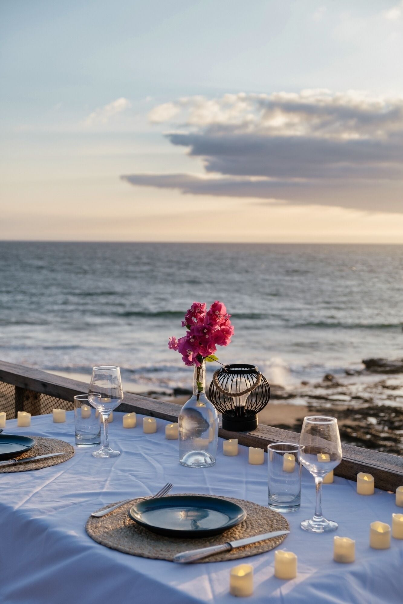 Mesa para dos con vista al océano, velas y un florero con flores rosadas.
