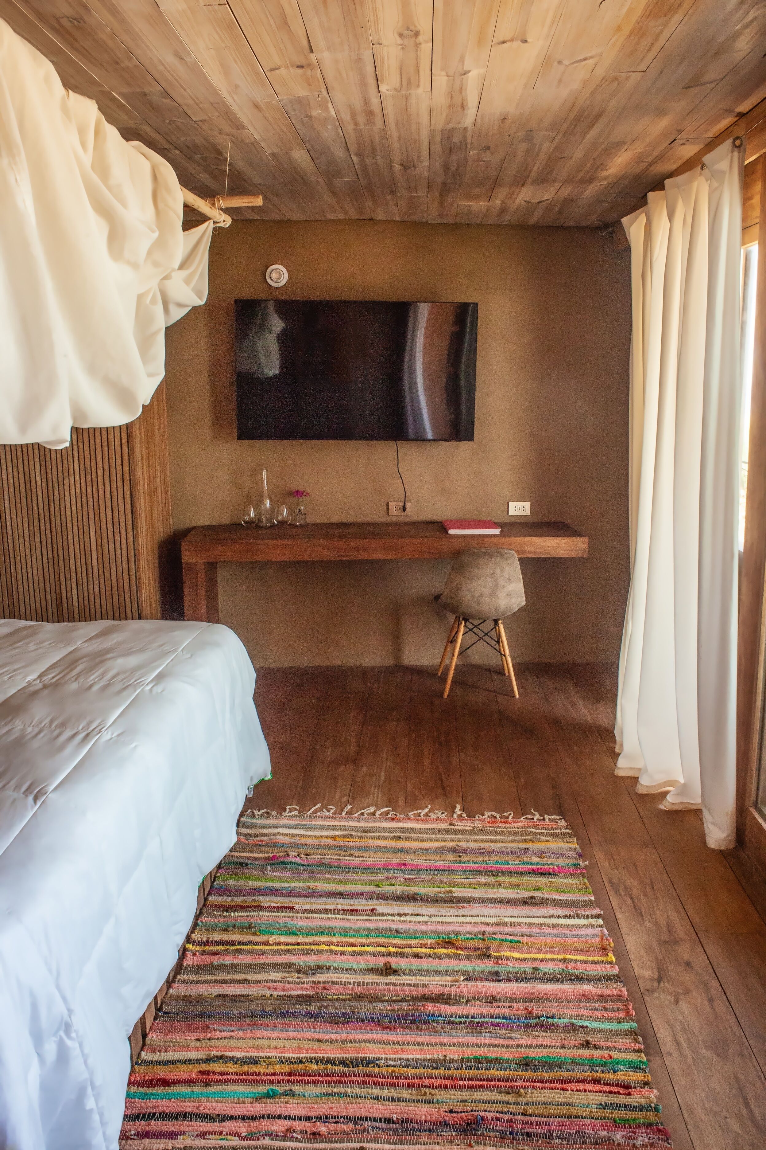 A bedroom with a bed, a desk, a chair, a hanging light, and a window with curtains.