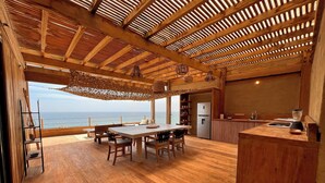 Una habitación amplia con piso de madera, una zona de comedor con mesa y sillas, un área de cocina con refrigerador y vista al mar.