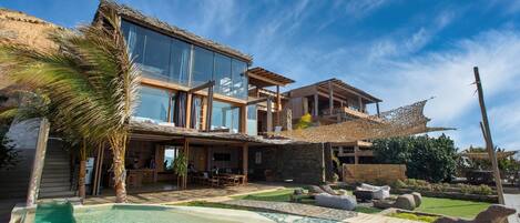 Una casa moderna con piscina, mobiliario exterior y una estructura de techo de paja.