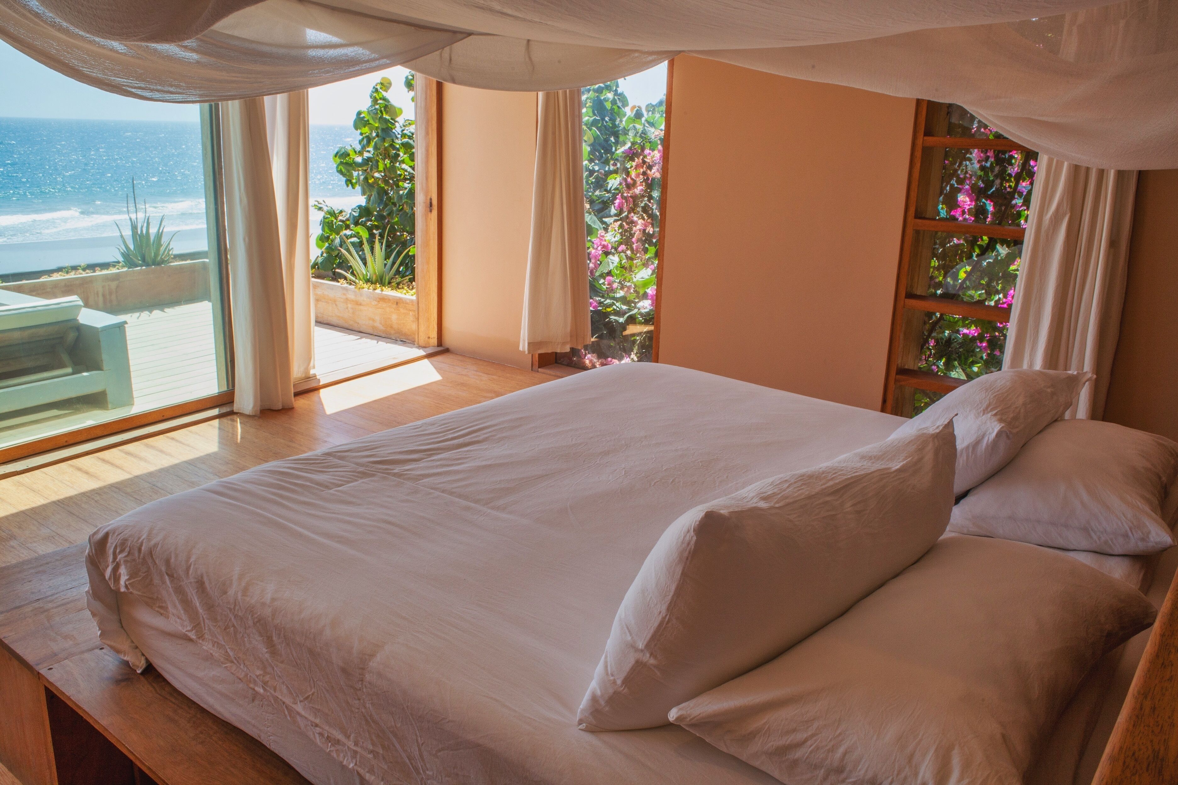 A bedroom with a large bed, a view of the ocean, and a balcony with plants.