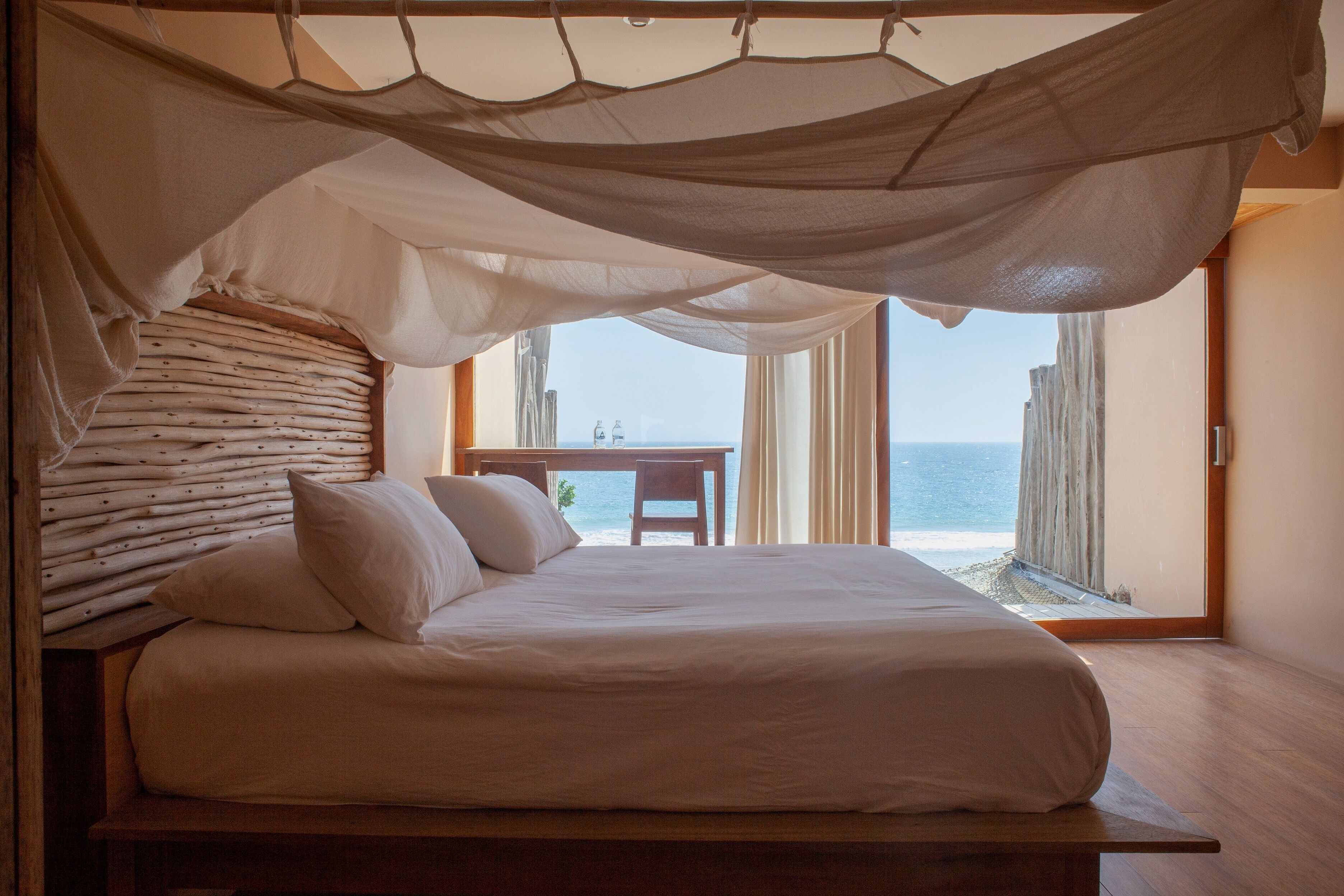 A bedroom with a bed, a wooden headboard, a table, and a view of the sea through large windows.