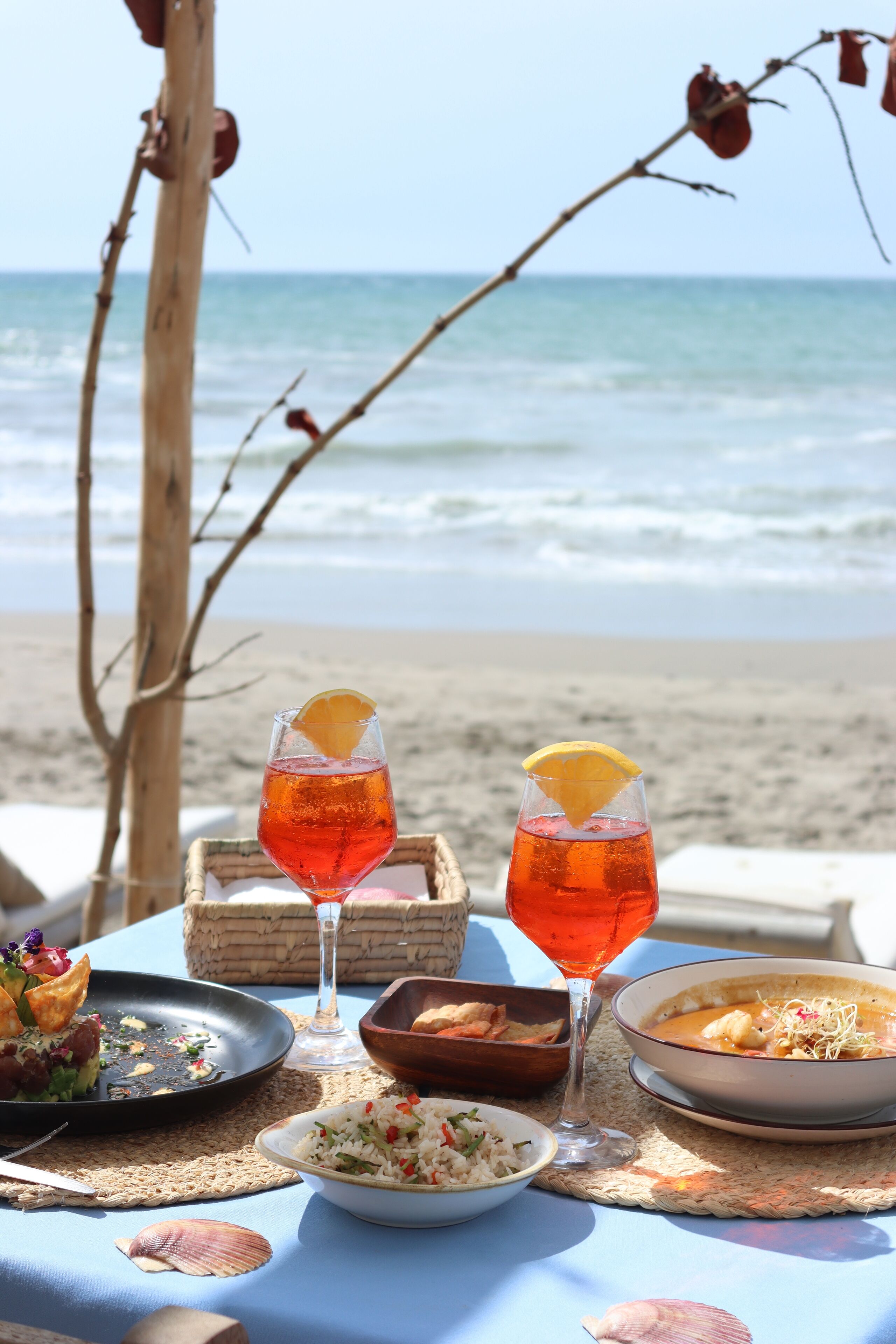 A table set with a meal and drinks on a beach with the ocean in the background.