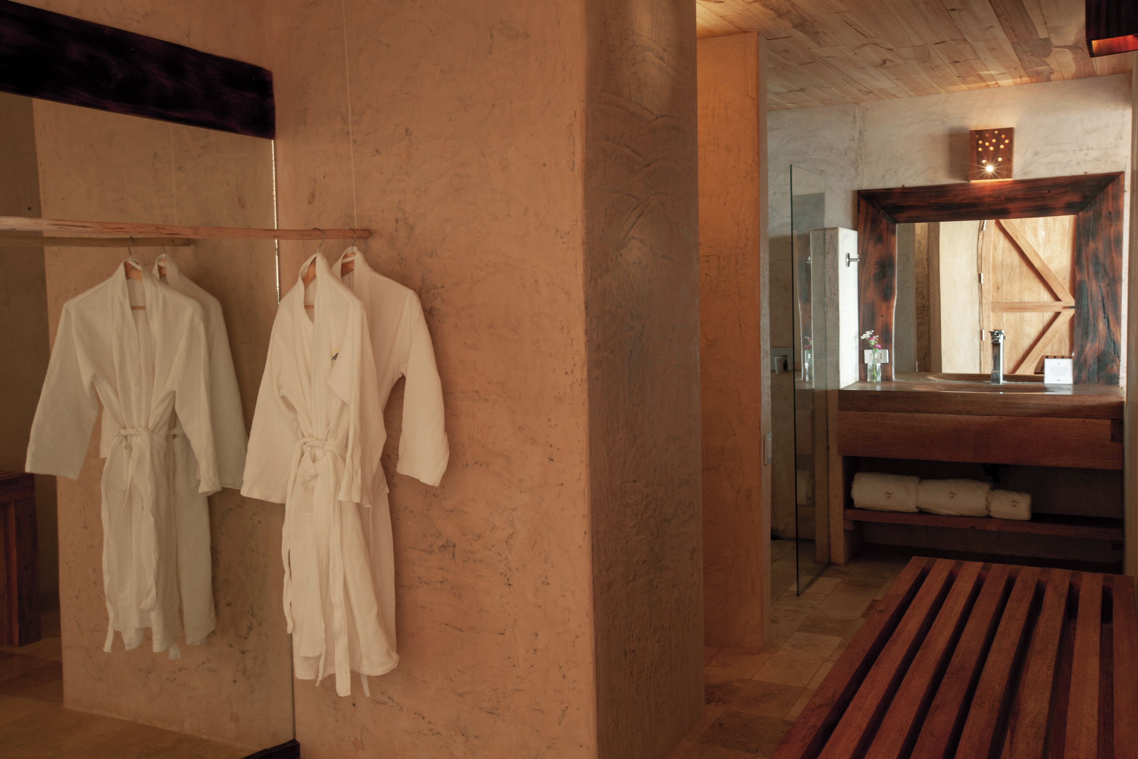 A bathroom with wooden benches, a mirror, and two white robes hanging on the wall.