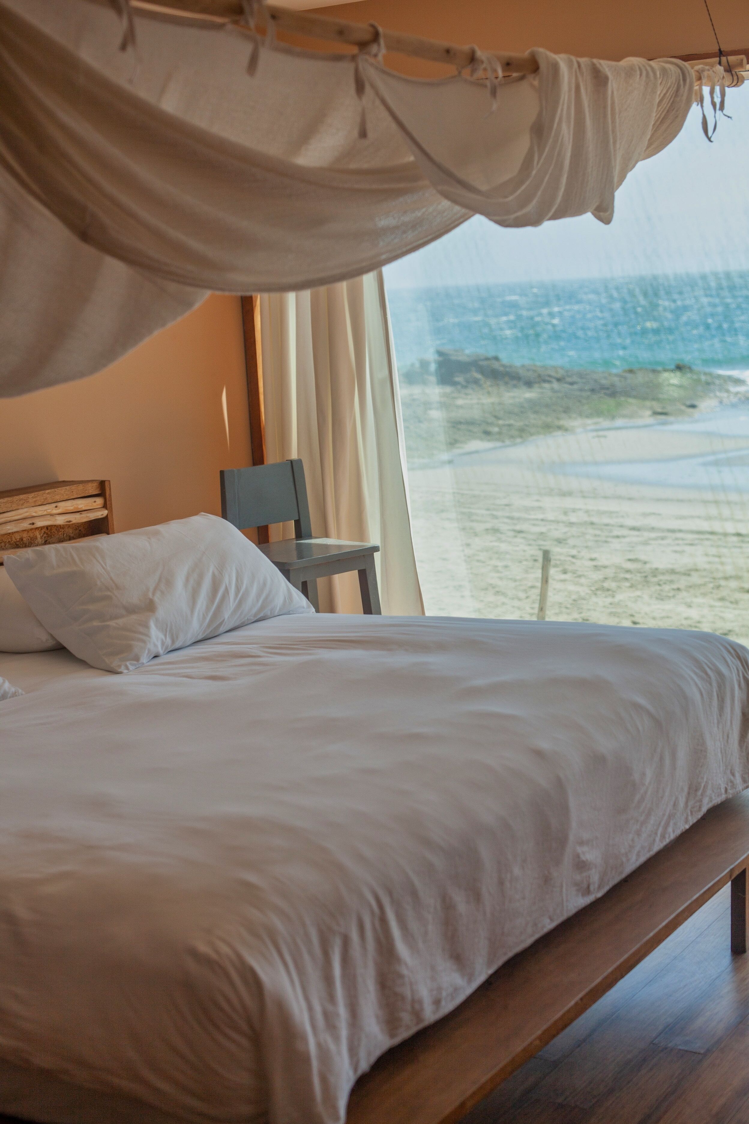 A bed with a canopy, a chair, and a view of the beach through a window.