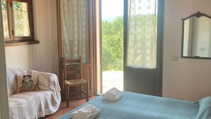 Une chambre à coucher avec un lit, une chaise et un canapé. Il y a une fenêtre avec des rideaux et une vue sur la verdure à l’extérieur.