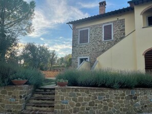 A stone house with a stone staircase leading to a door, surrounded by vegetation and a clear sky.