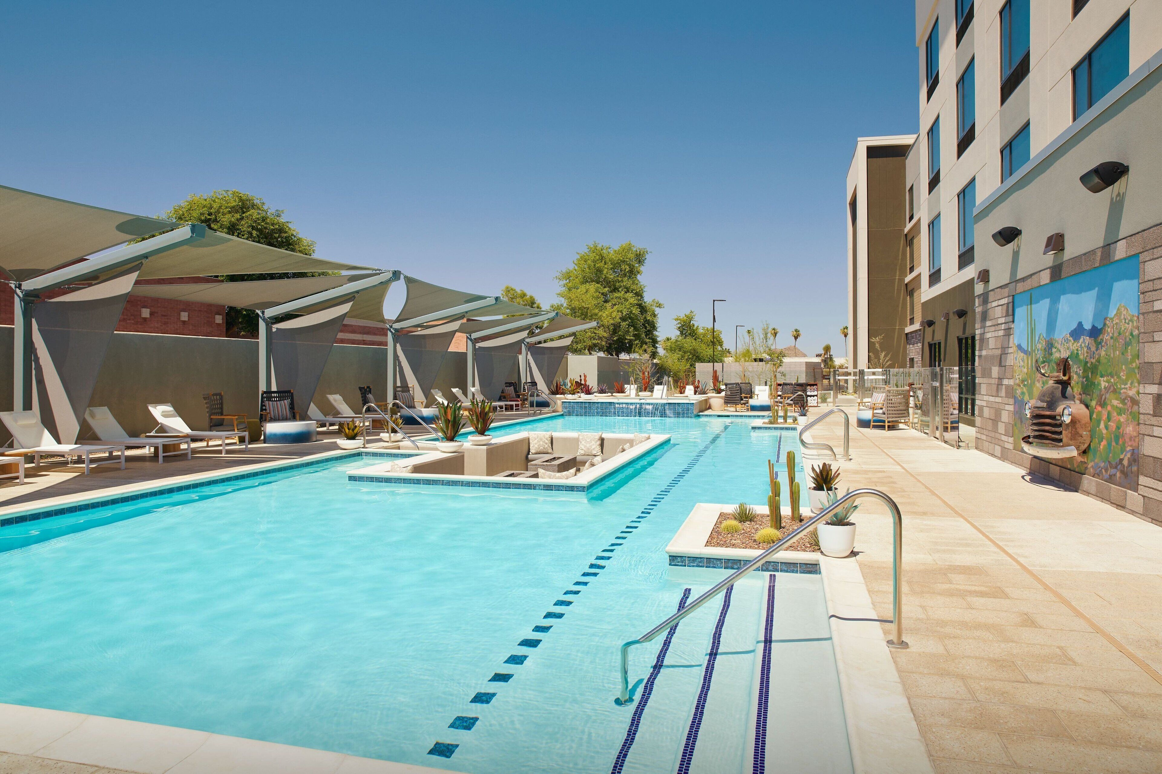 Piscina com espreguiçadeiras e guarda-sóis, cercada por um edifício com um mural.