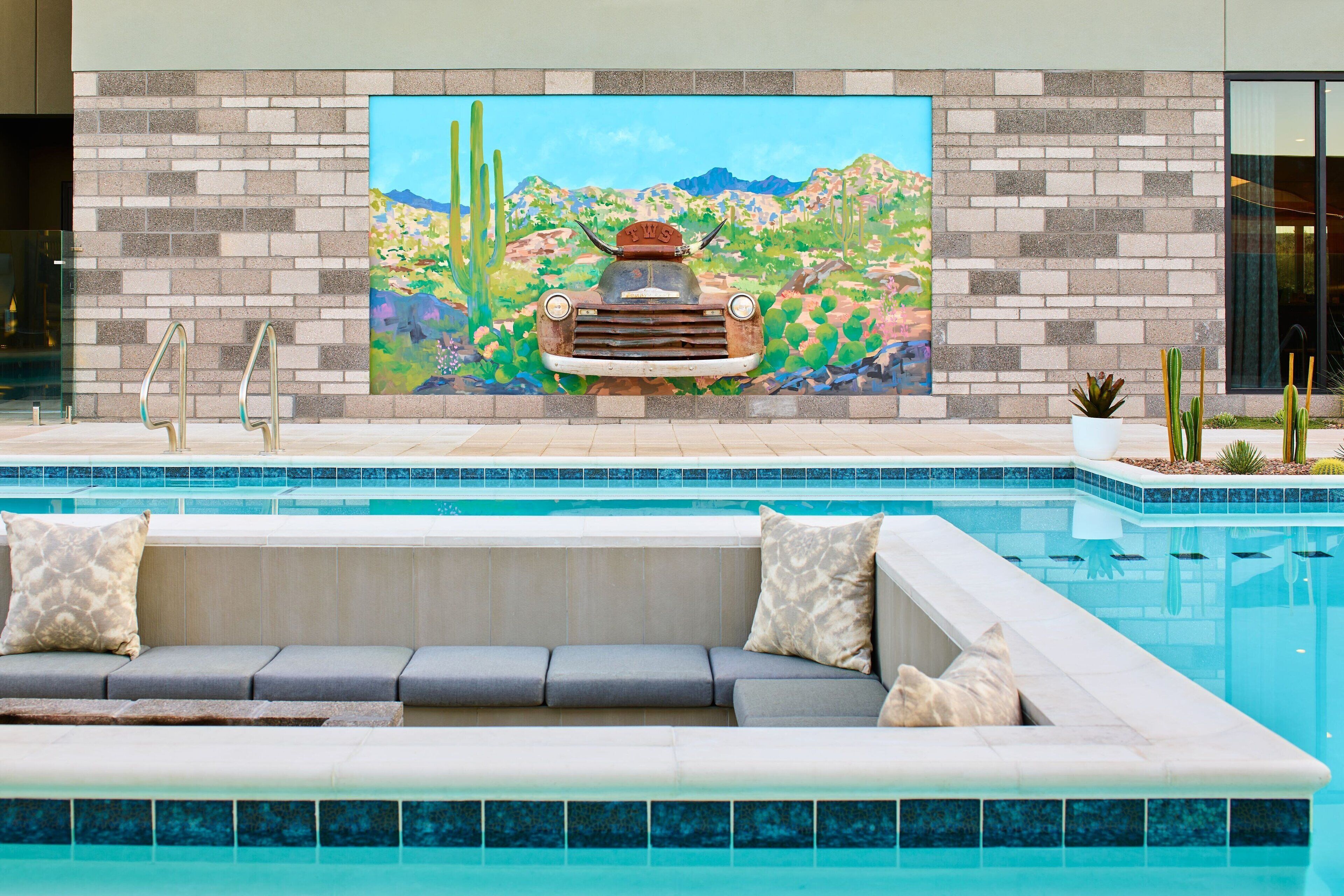 Et moderne poolområde med et muralmaleri af en bil og et ørkenlandskab, siddepladser og frit udsyn til poolen.