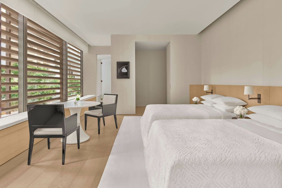 Habitación de hotel con dos camas, un escritorio con silla, una mesita y una ventana con persianas.