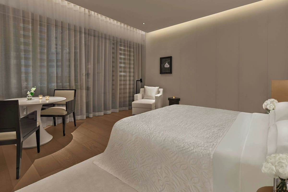 Una habitación de hotel moderna con una cama grande, un escritorio con sillas, una butaca, una mesita con flores y un cuadro en la pared.