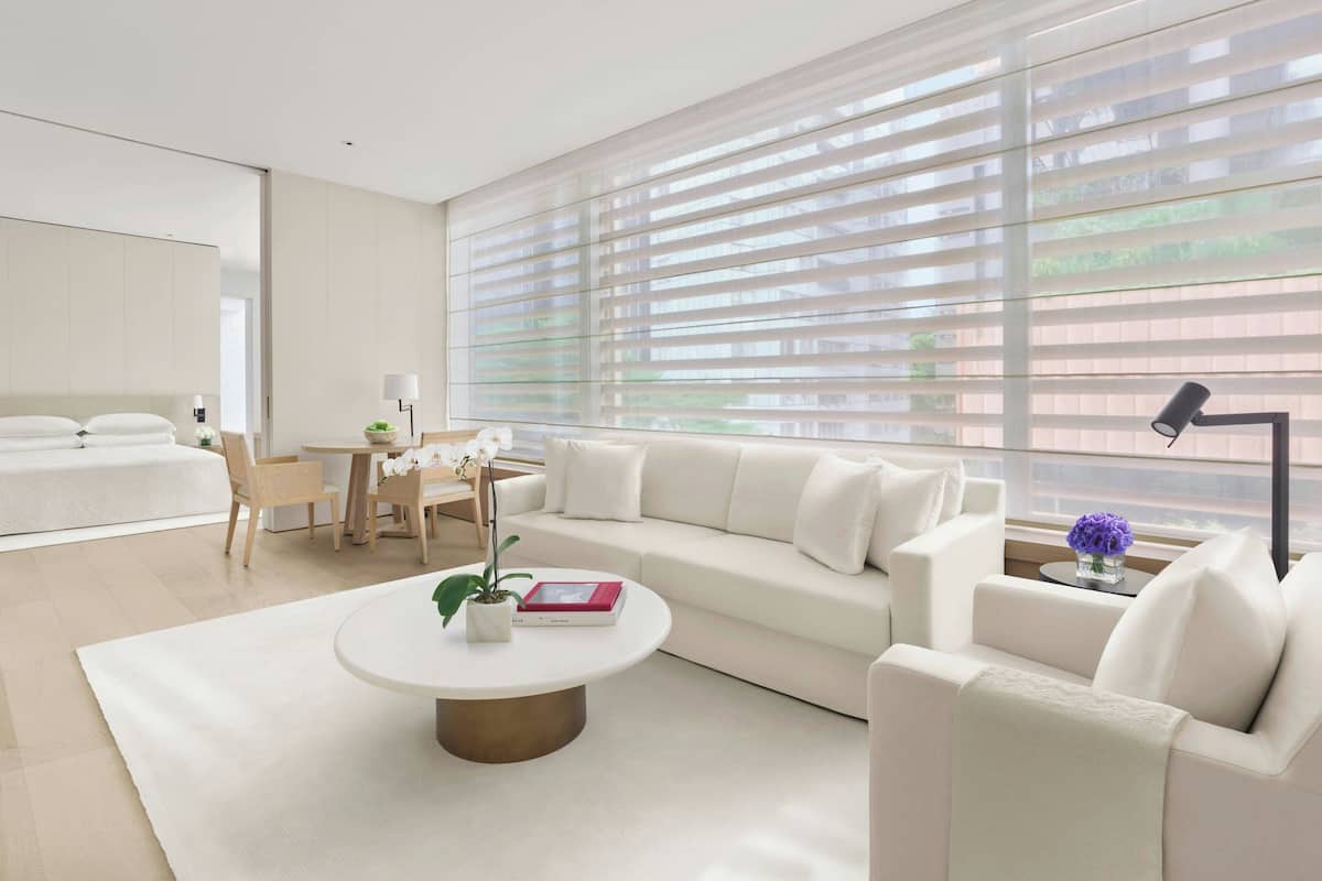 Una habitación de hotel moderna con un ventanal grande, un sofá blanco, una mesa redonda con una planta, una mesita con una lámpara y una cama con ropa de cama blanca.
