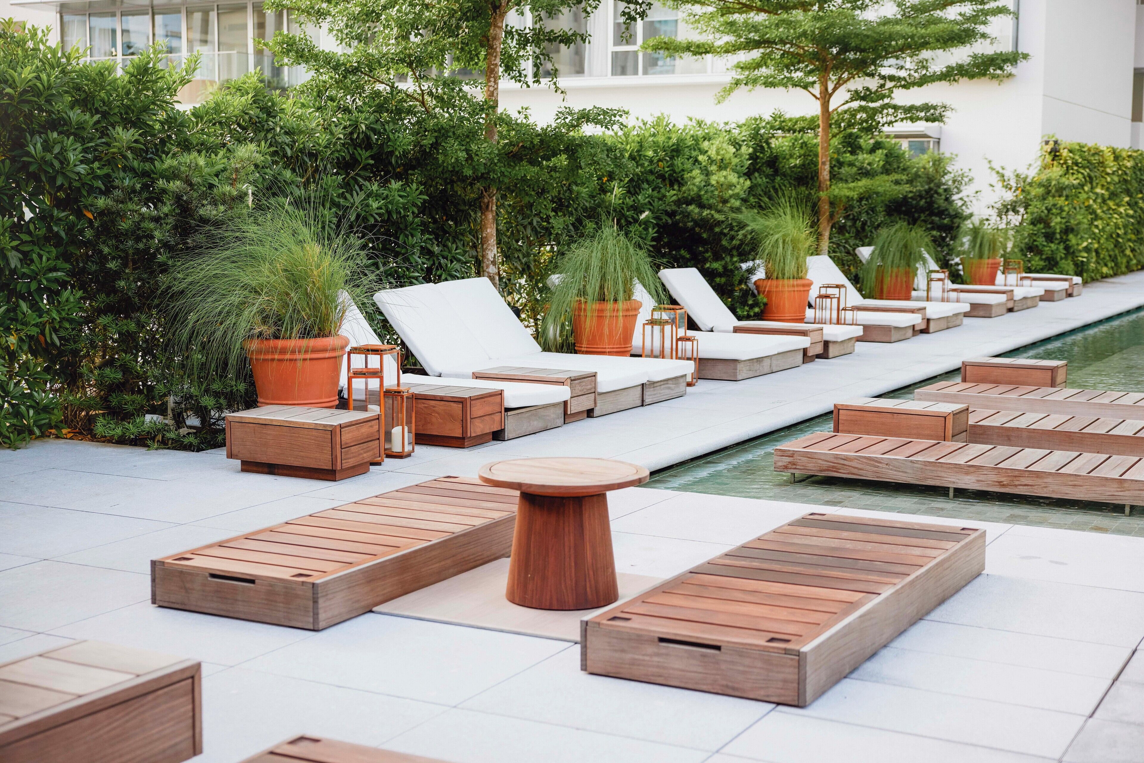 Un área junto a la piscina con tumbonas de madera, cojines blancos y plantas en macetas.