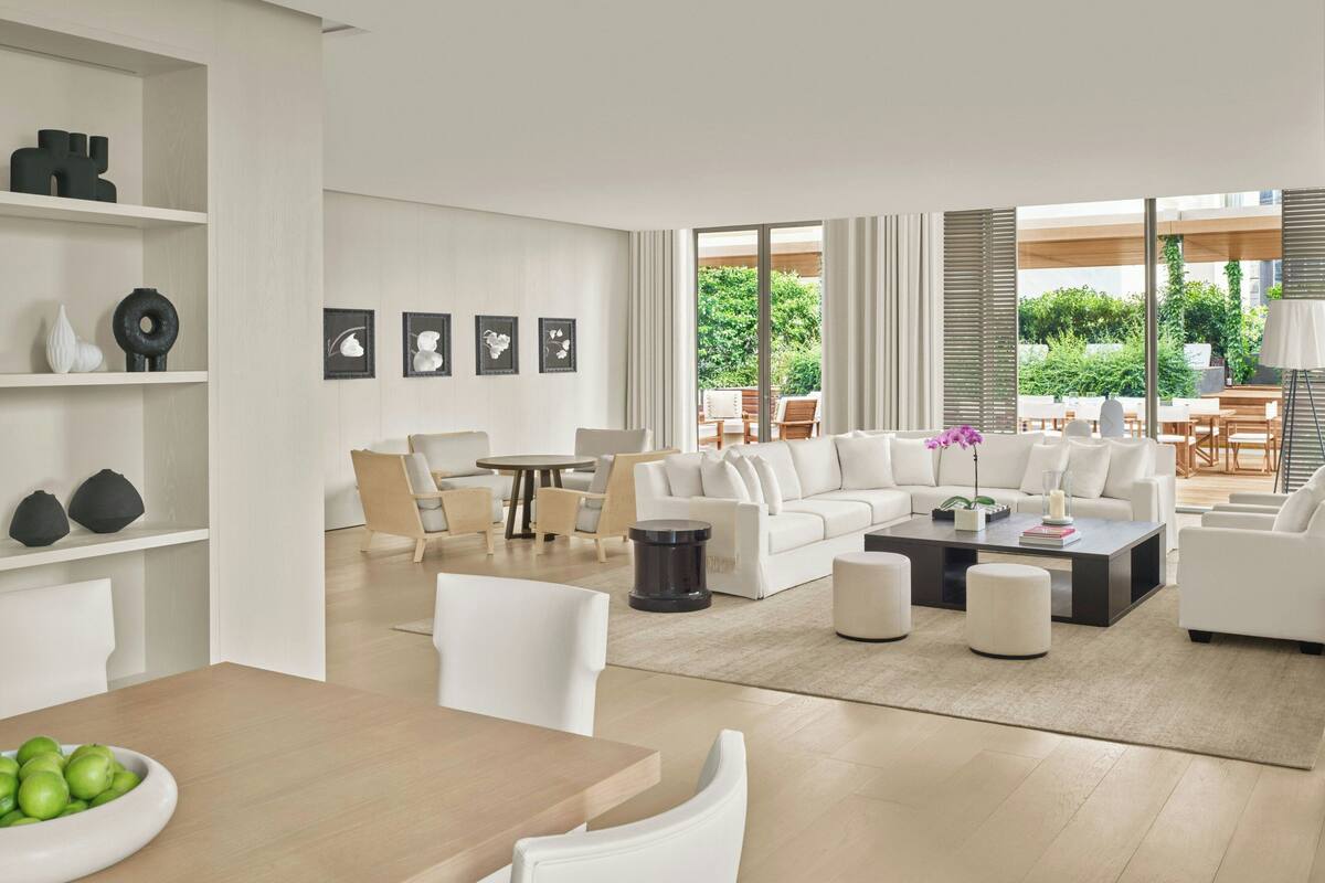 Una sala de estar moderna con un sofá modular, una mesa de centro y una zona de comedor con una mesa y sillas. Hay cuadros enmarcados en la pared, y vistas al exterior a través de grandes ventanales.