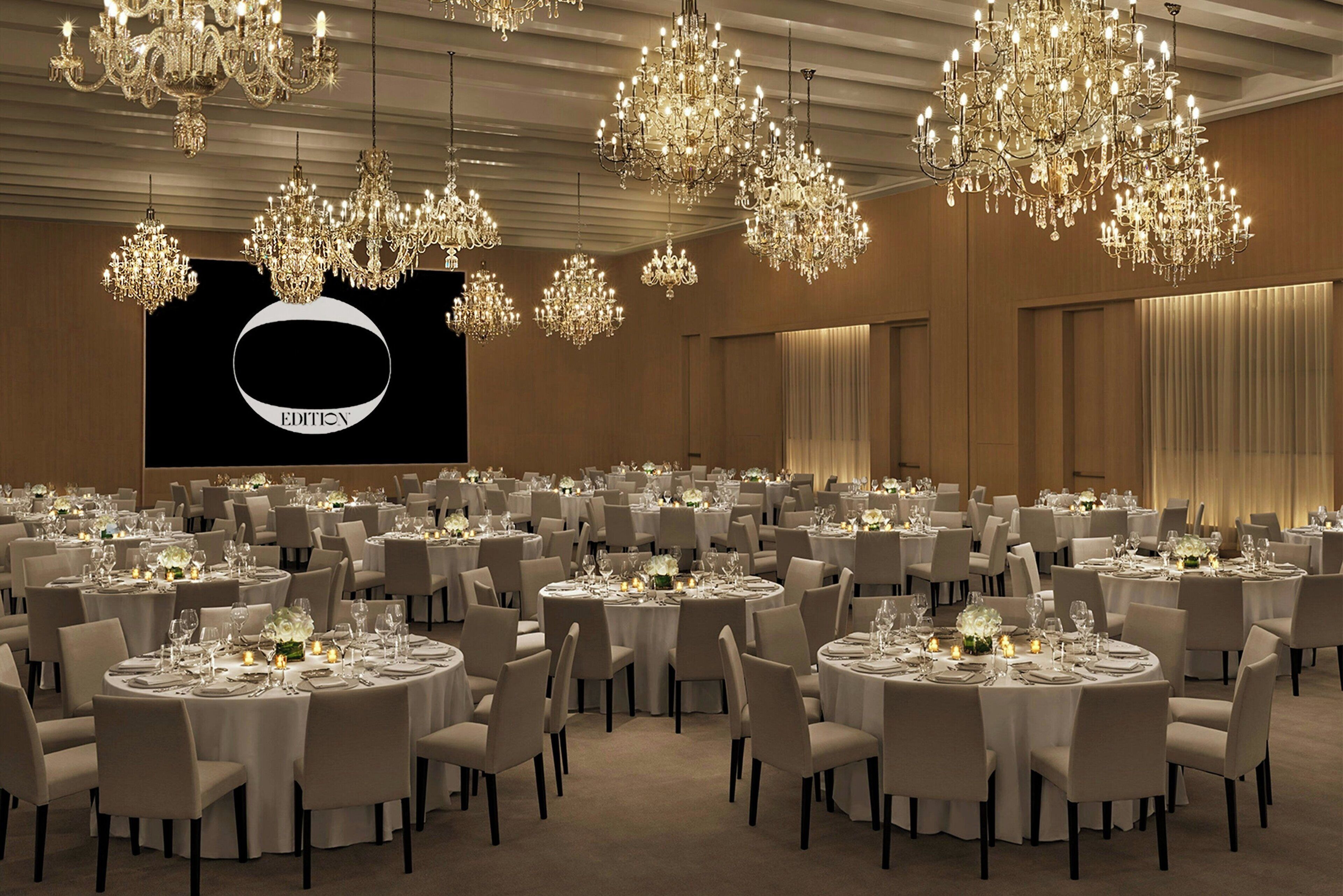 A banquet hall with multiple round tables, each set with white tablecloths and chairs, and ornate chandeliers hanging from the ceiling.
