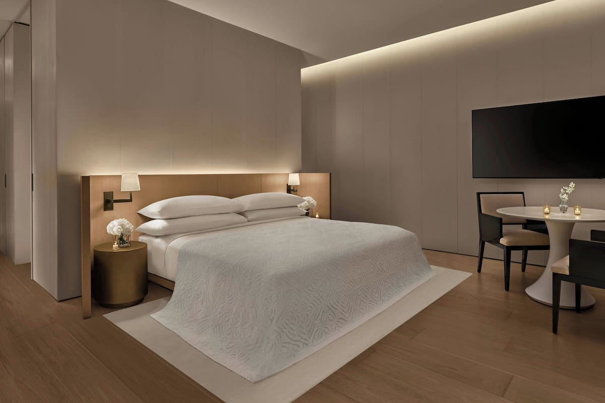 Una habitación de hotel moderna con una cama grande, lámparas de noche, una mesa redonda con sillas y un televisor de pantalla plana montado en la pared.