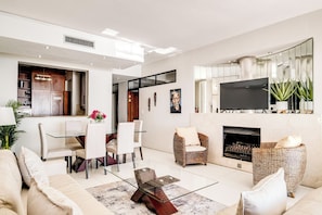 A modern living room with a glass coffee table, a fireplace, and a dining area with white chairs.