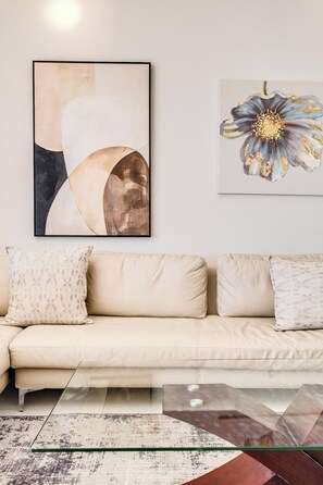 A modern living room with a beige sofa, two patterned throw pillows, a glass coffee table, and two abstract paintings on the wall.