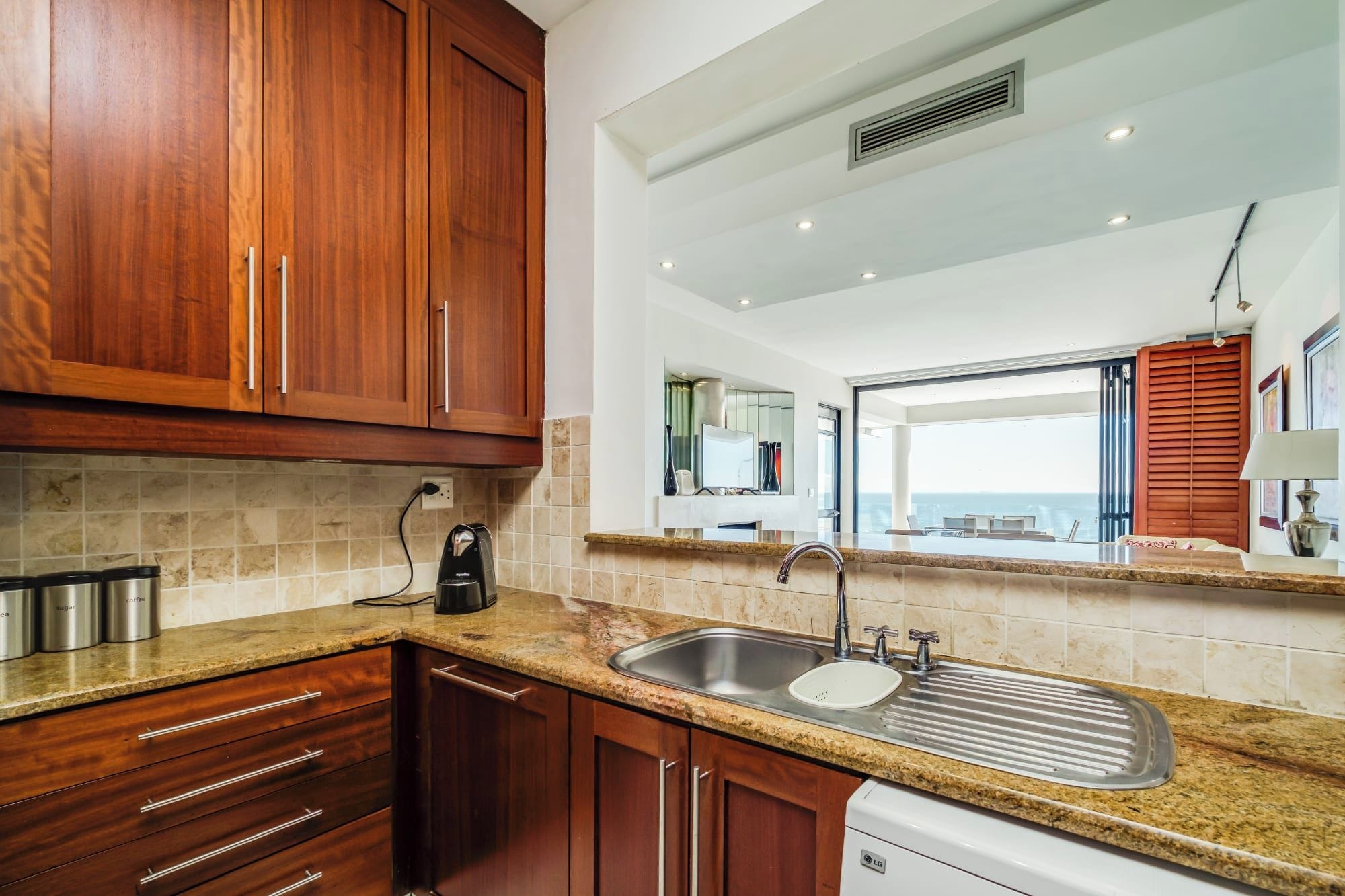 A modern kitchen with wooden cabinets, a granite countertop, stainless steel appliances, and a view of the sea through a large window.