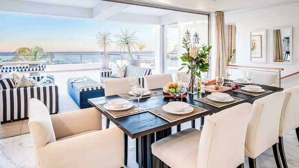 A dining area with a view of the ocean, featuring a dark wooden table set with white plates and glasses, surrounded by beige upholstered chairs. - Immaculate Camps Bay Villa w Jacuzzi, Views and Pool Villa Grenache (Cape Town)