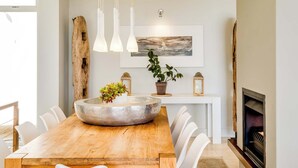 Uma sala de jantar com mesa de madeira, cadeiras brancas, uma grande tigela decorativa, luminárias pendentes, um vaso com planta, lareira e um quadro na parede.