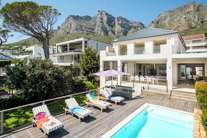 A modern two-story house with a swimming pool, lounge chairs, and a view of mountains.