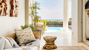 Uma sala de estar moderna com vista para uma piscina e para o mar, apresentando um sofá, mobiliário de vime e almofadas decorativas.