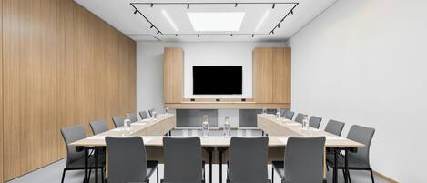 Uma sala de conferências com uma mesa comprida, cadeiras cinzentas, uma TV de tela plana e painéis de madeira em uma parede.