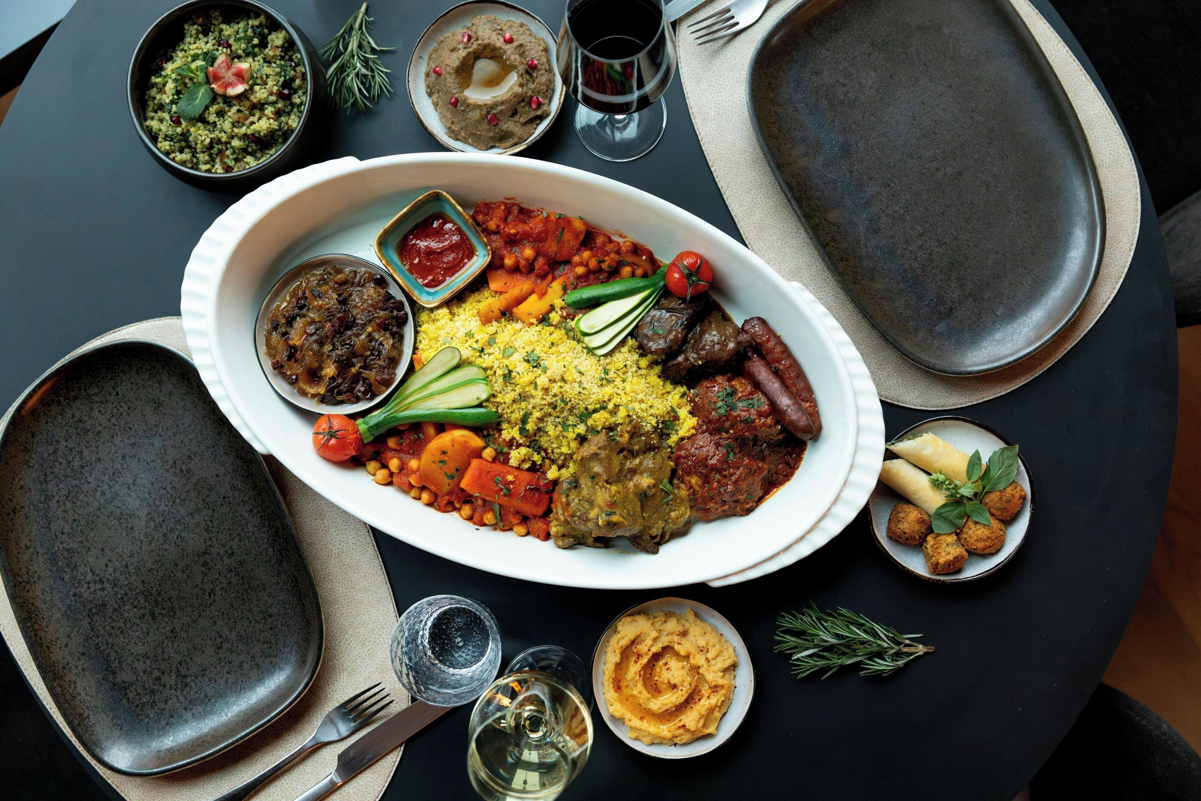 A table set with a variety of dishes, including a large plate of couscous, vegetables, and meat, accompanied by small bowls of dips and sauces.