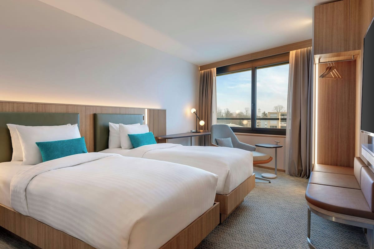 A hotel room with two beds, a desk, a chair, and a view of the city.