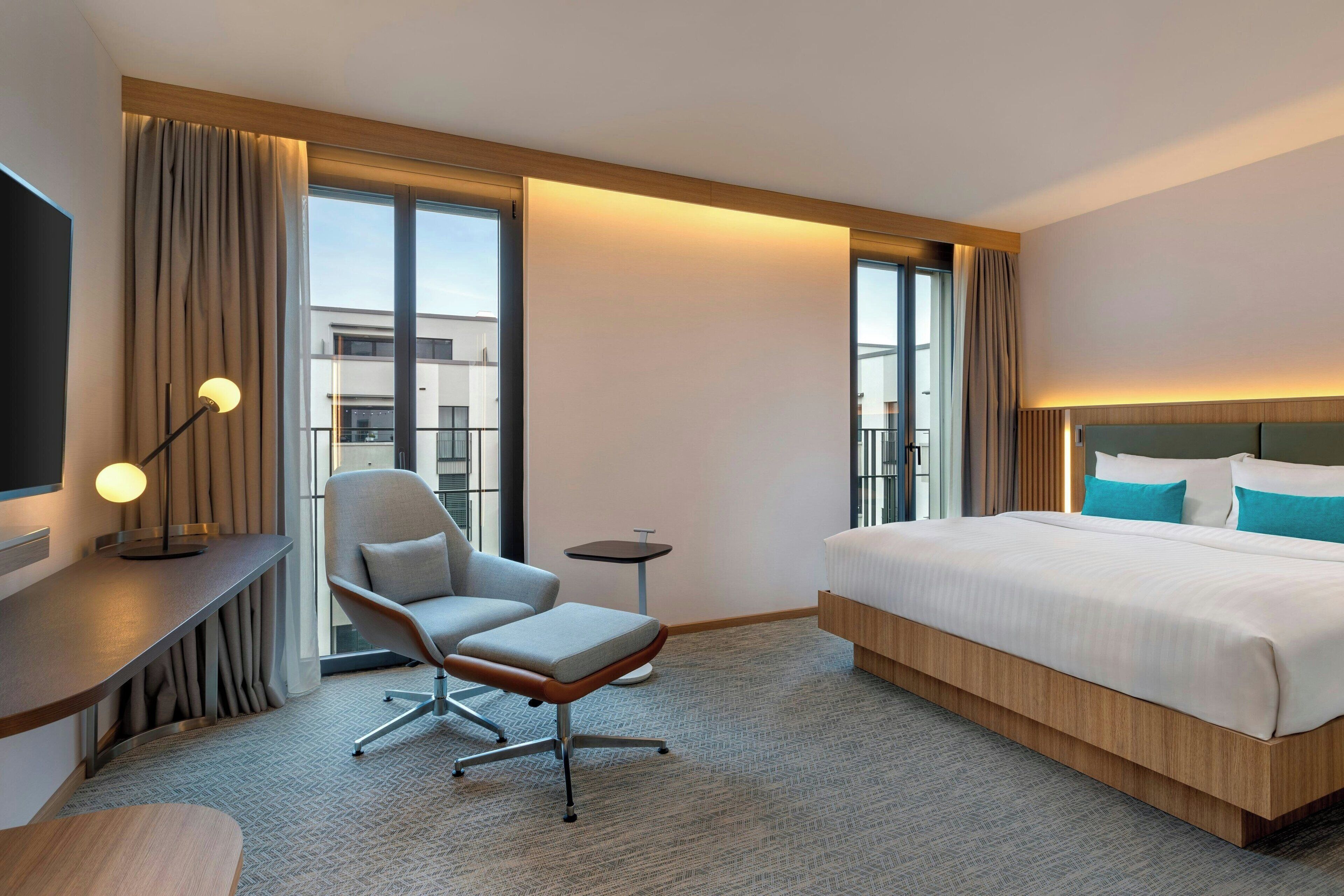 A hotel room with a bed, a desk, a chair, a TV, and large windows.