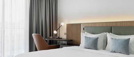 Habitación de hotel con una cama grande, un escritorio con lámpara y vistas al exterior a través de cortinas translúcidas.