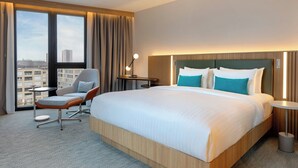 Habitación de hotel con una cama grande, una silla, un escritorio y vistas a la ciudad a través de amplias ventanas.