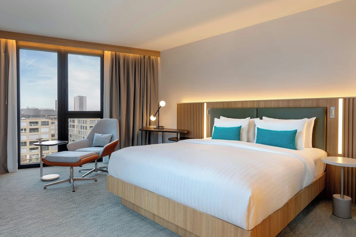 A hotel room with a large bed, a chair, a desk, and a view of the city through large windows.