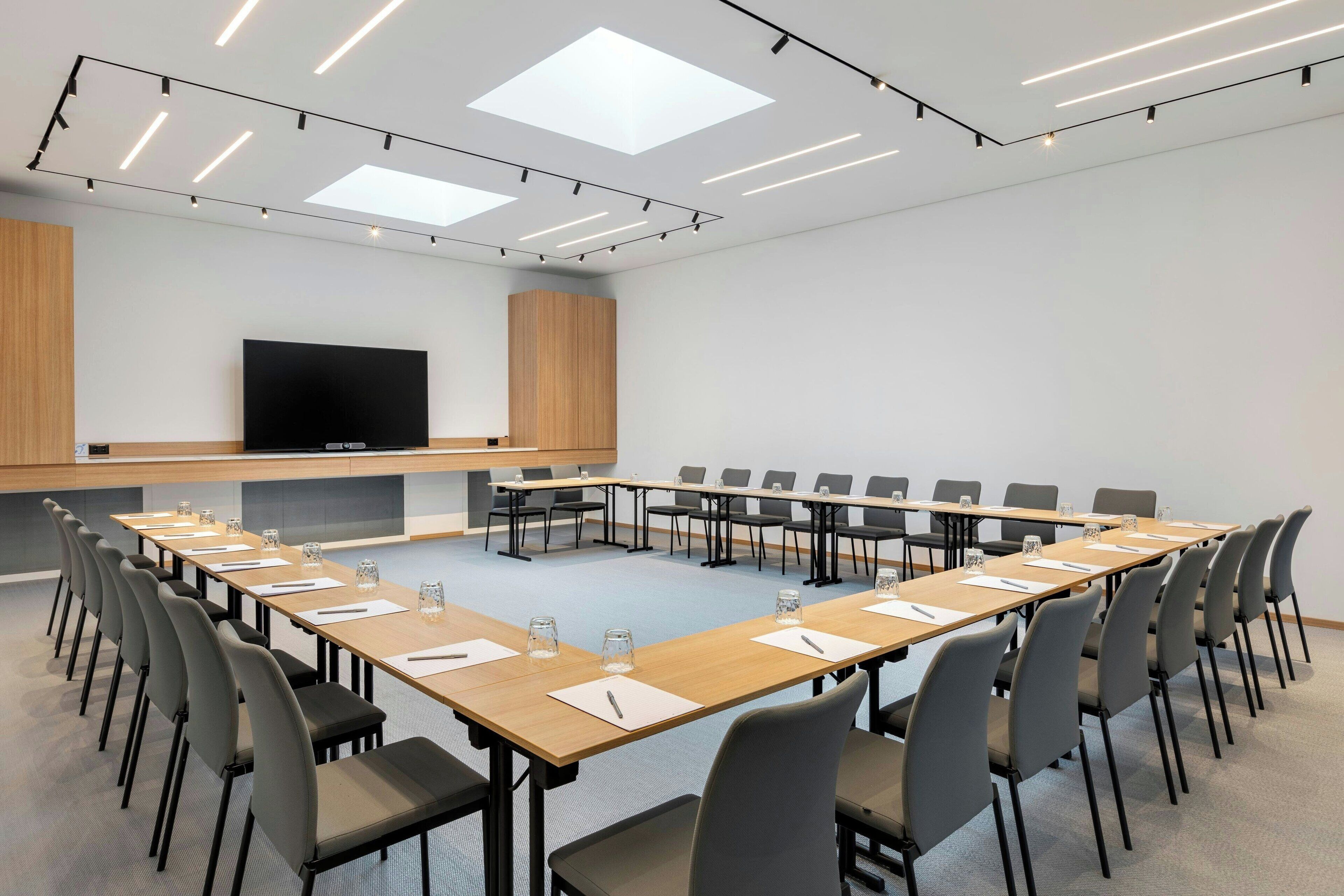 Un salón de conferencias con una larga mesa, sillas, un televisor de pantalla plana y luces en el techo.